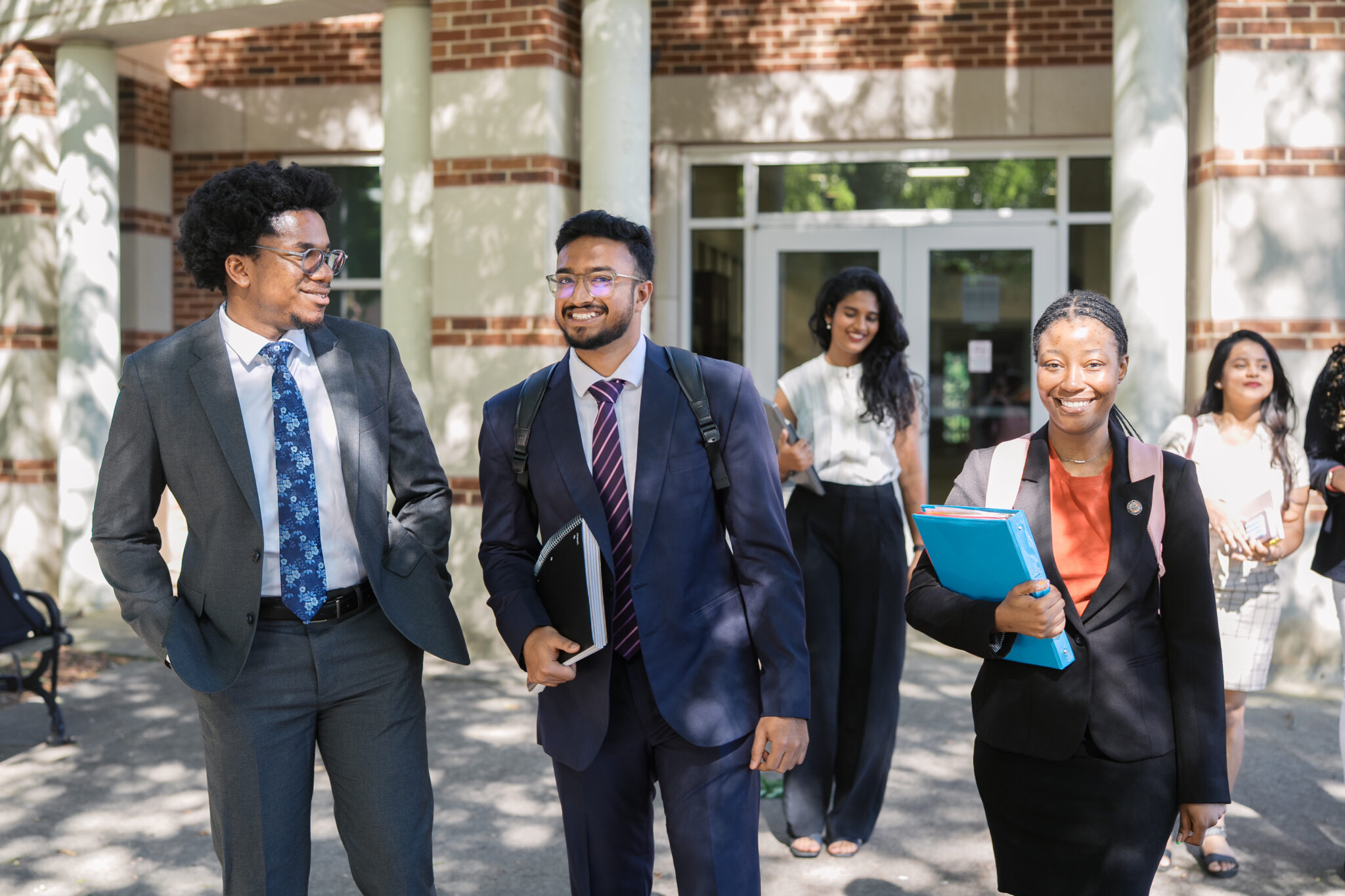 Three young professionals in business attire walk outside a building, holding folders and smiling, with others in the background.