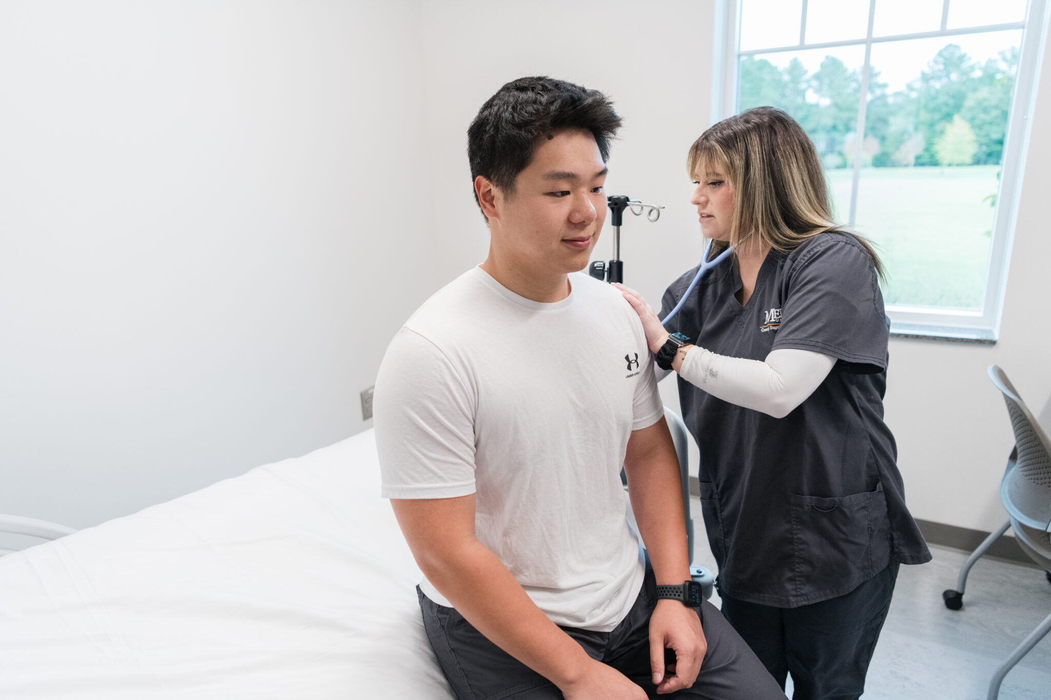 A nursing student uses a stethoscope to examine a seated patient in a medical exam room.