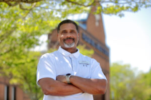 A man stands with arms crossed in front of a brick building.