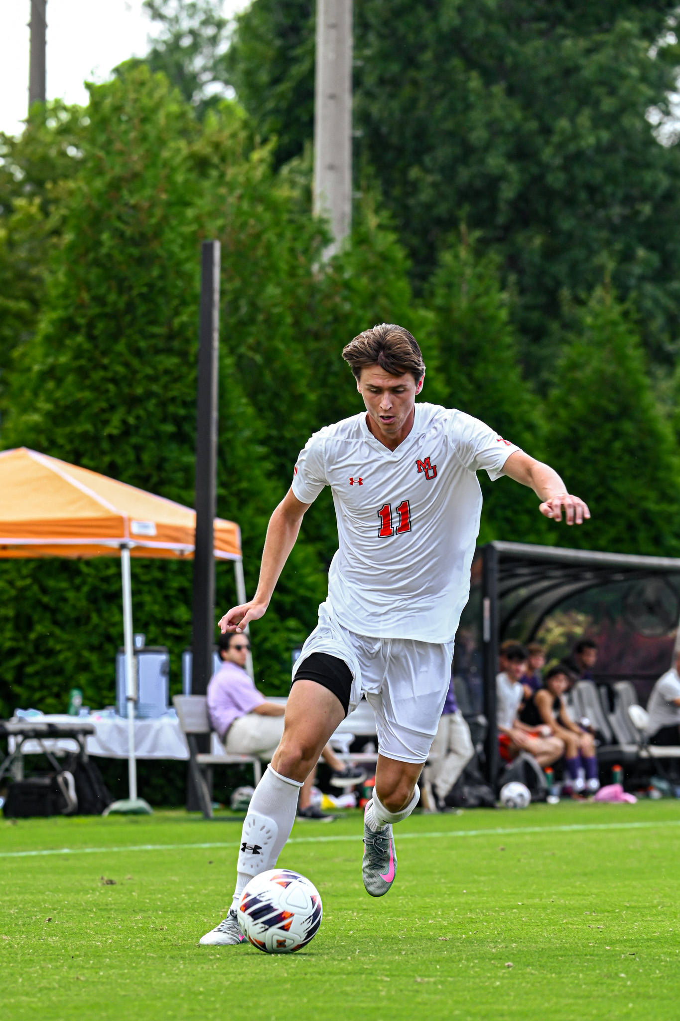 A Mercer men's soccer player dribbles the ball downfield with players and a tent in the background.