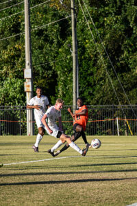 A Mercer men's soccer player battles for the ball against two opponents during a sunny match.
