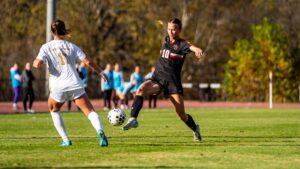 A Mercer women's soccer player kicks the ball mid-air while an opponent defends on the field.