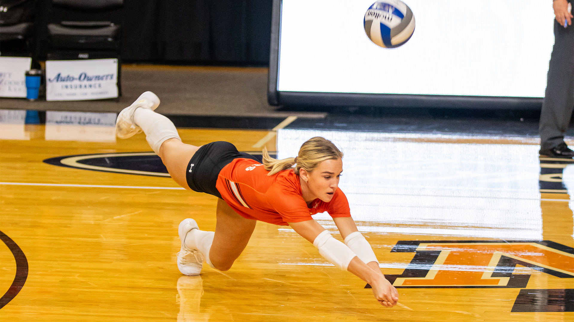 A Mercer volleyball player dives parallel to the floor, fully extended, to dig a ball just out of reach during a match on an indoor wood court.