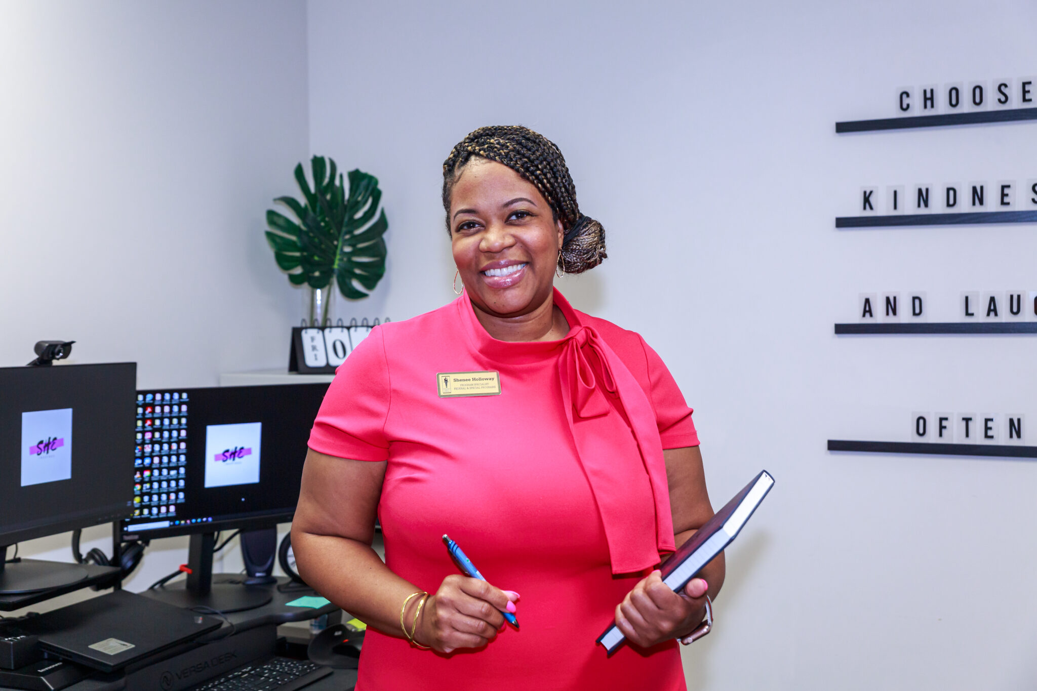 A smiling woman in a pink dress holds a notebook and pen in an office, with a Choose Kindness wall sign and dual monitors behind her.
