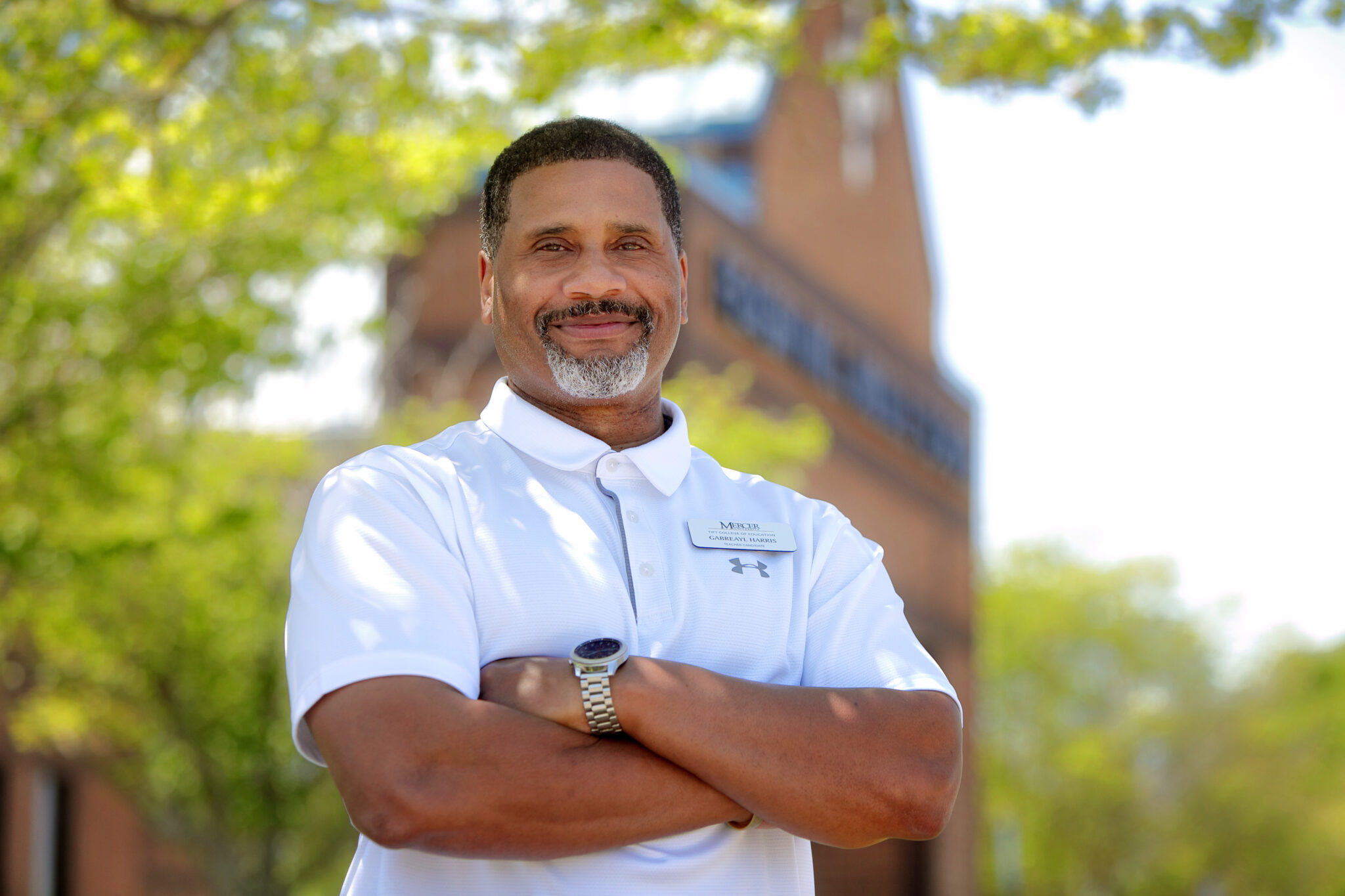 A man stands with arms crossed in front of a brick building.