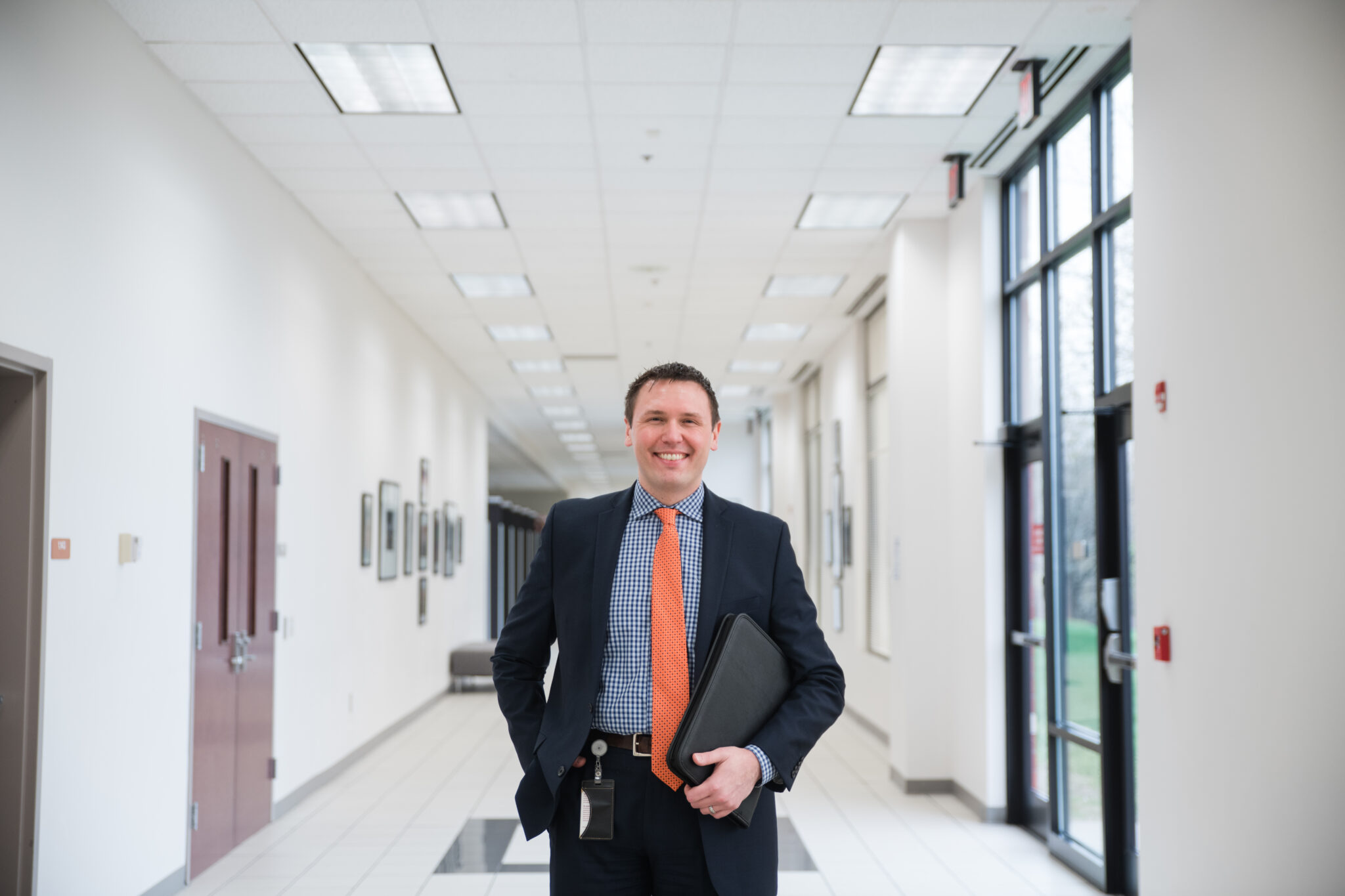 A smiling man in a navy suit and orange tie holds a portfolio while standing in a bright institutional hallway.