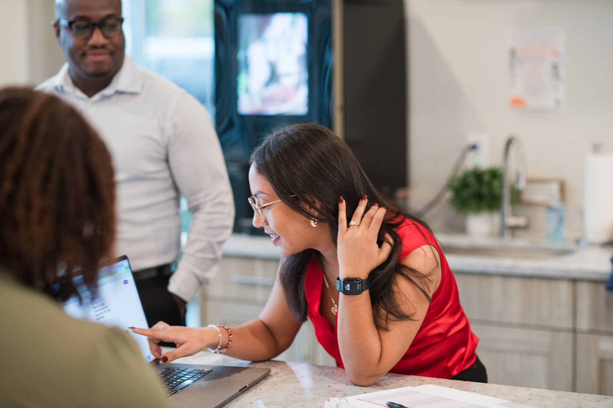 A woman smiles while pointing at a laptop screen during a collaborative discussion with two colleagues in a bright workspace.