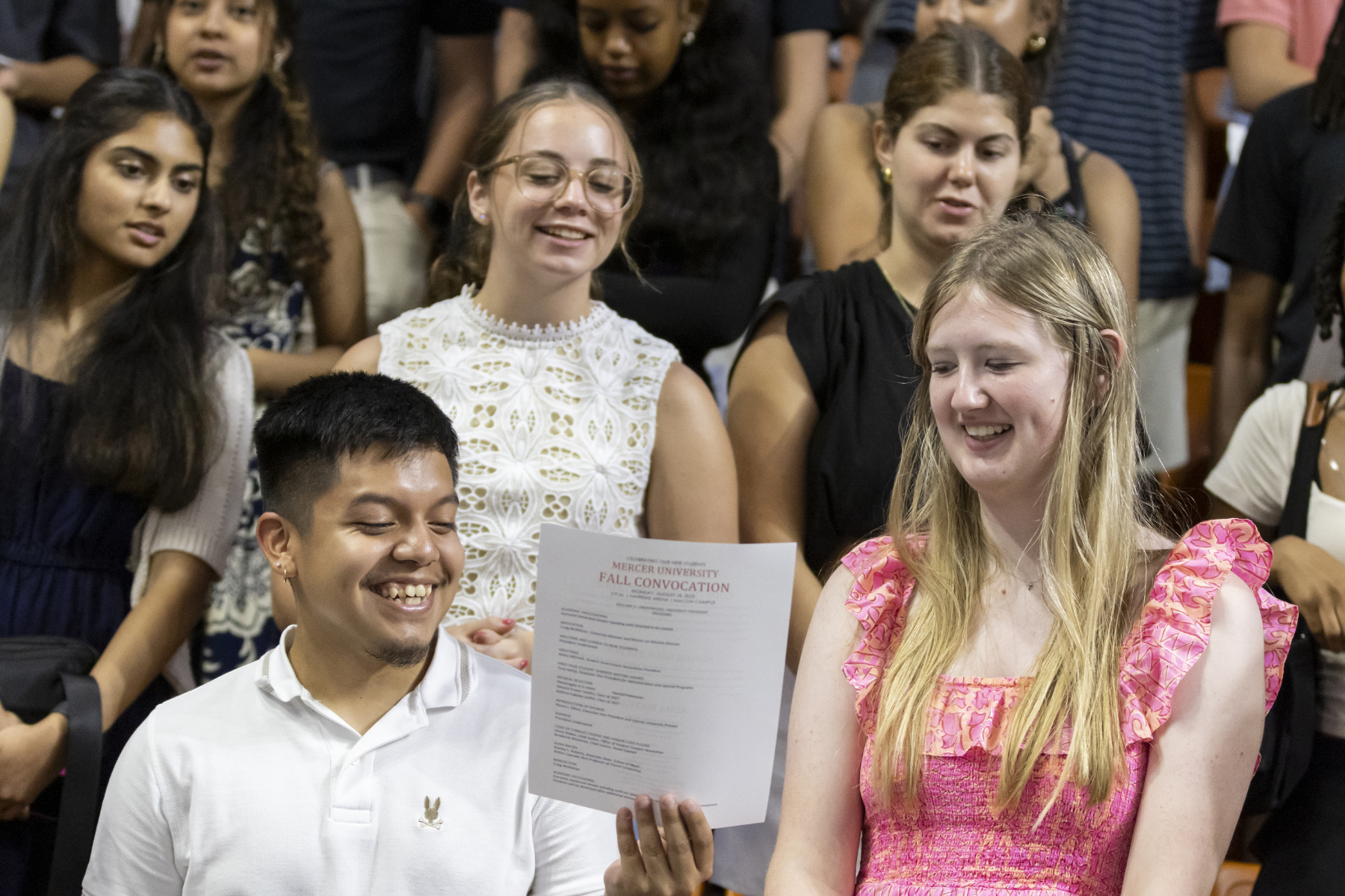 Two smiling Mercer University students share a Fall Convocation program while seated among fellow students at the ceremony.