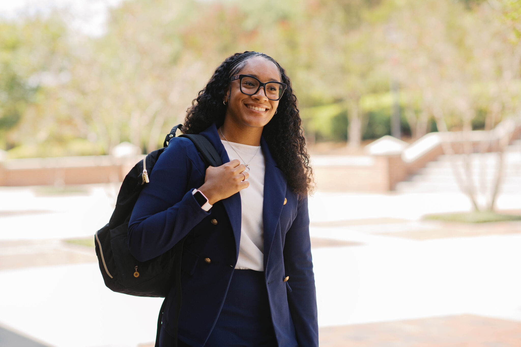 A woman in business attire smiles while carrying a backpack outdoors on a sunny day.