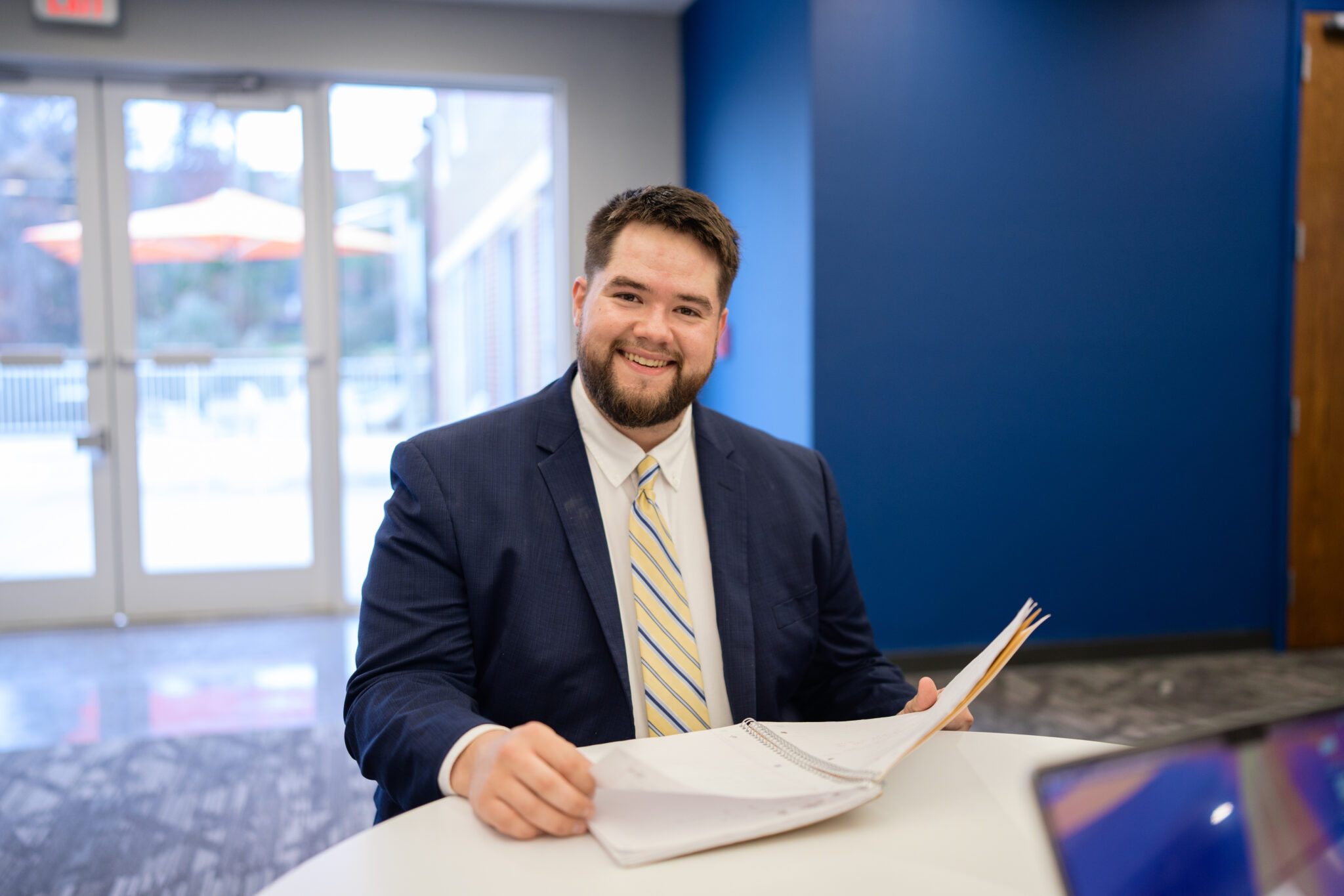 A smiling MBA student in a navy suit and striped tie looks up from an open notebook at a table in a campus building.
